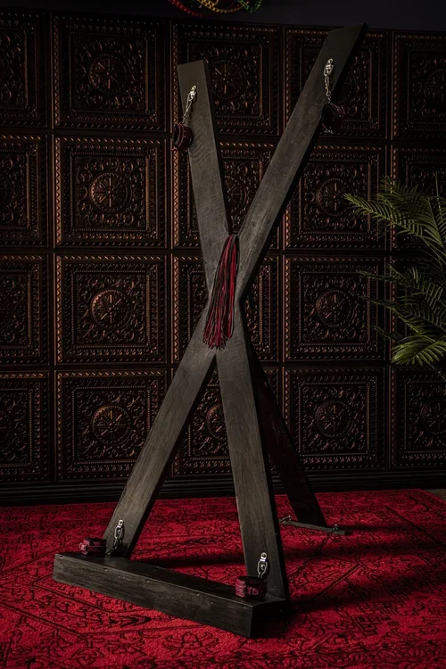 A large wooden X-shaped guillotine lock in front of an ornate carved wooden wall and red carpet, with a green plant on the right side.