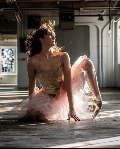 A ballerina in a pink tutu is sitting on the floor of a dance studio, looking to the side with her hair flowing and illuminated by natural light coming from large windows.