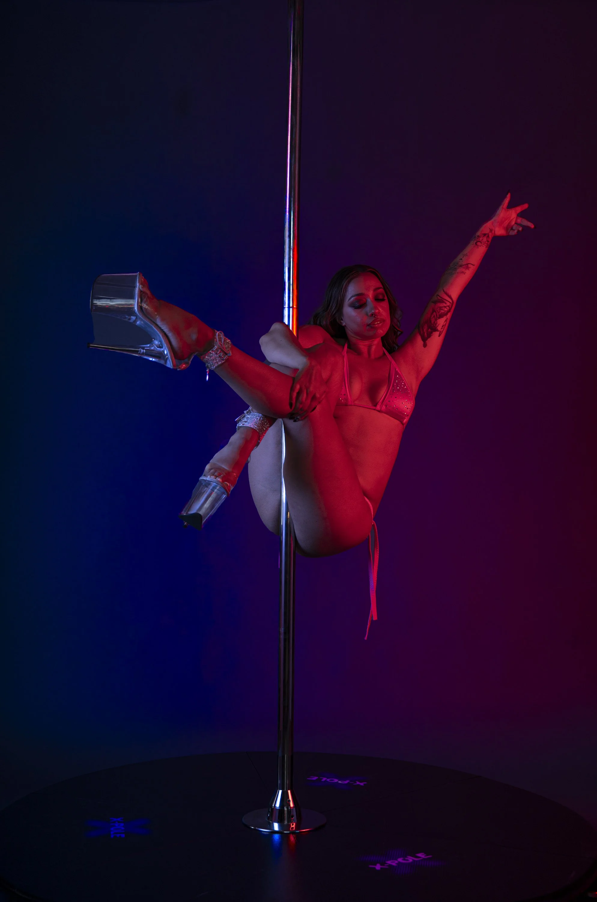 A woman performing a pole dance, holding a clear high-heeled shoe with rhinestones in front of a dark background with colored lights.