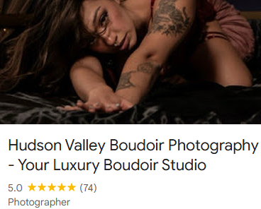 A woman with glasses lying on her side on a bed, resting her head on her arm, with tattoos on her arm.