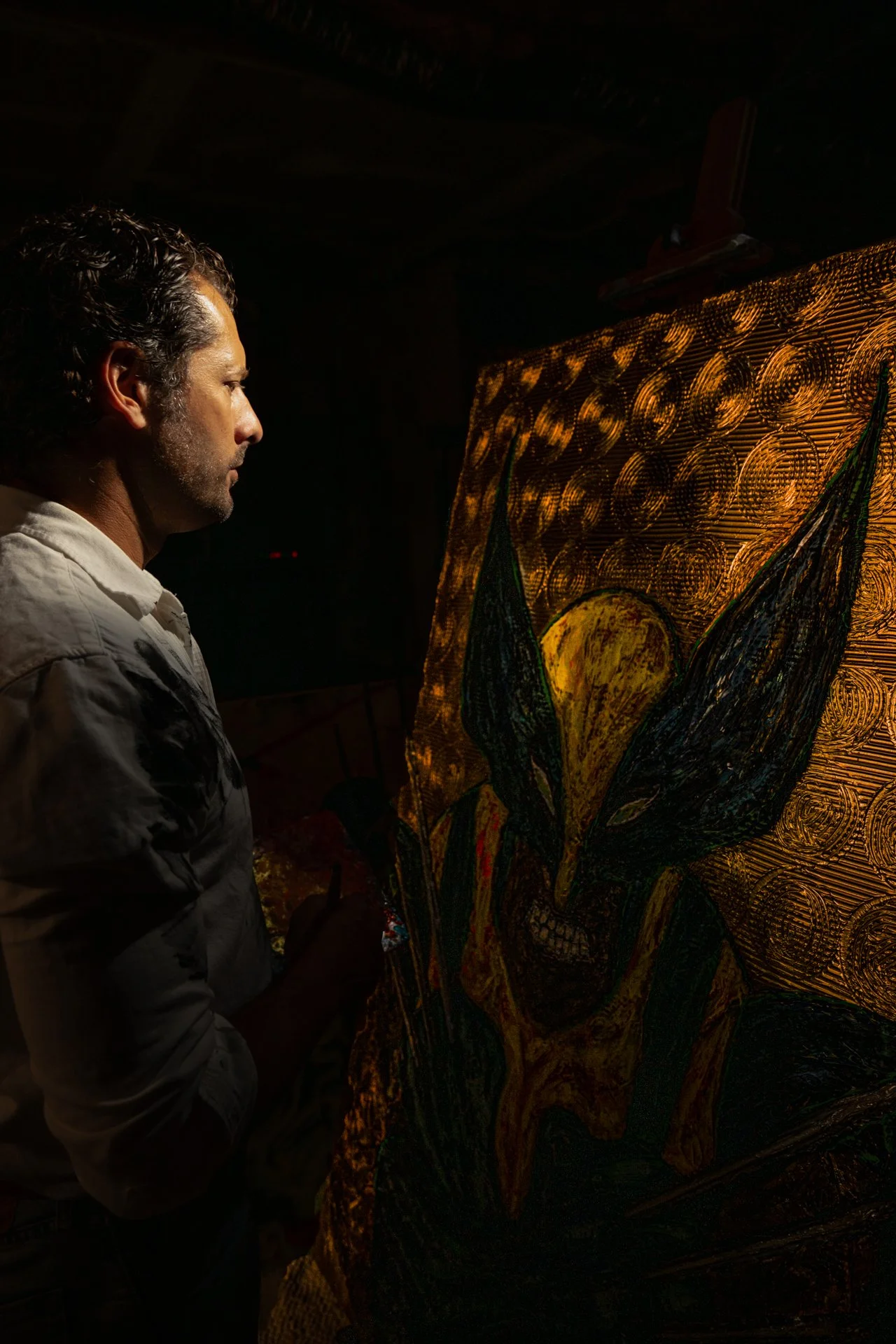 A man with dark, curly hair and a light-colored shirt stands in front of a brightly illuminated, colorful painting. The painting features a stylized flower with yellow and black petals and a background with numerous golden circles.