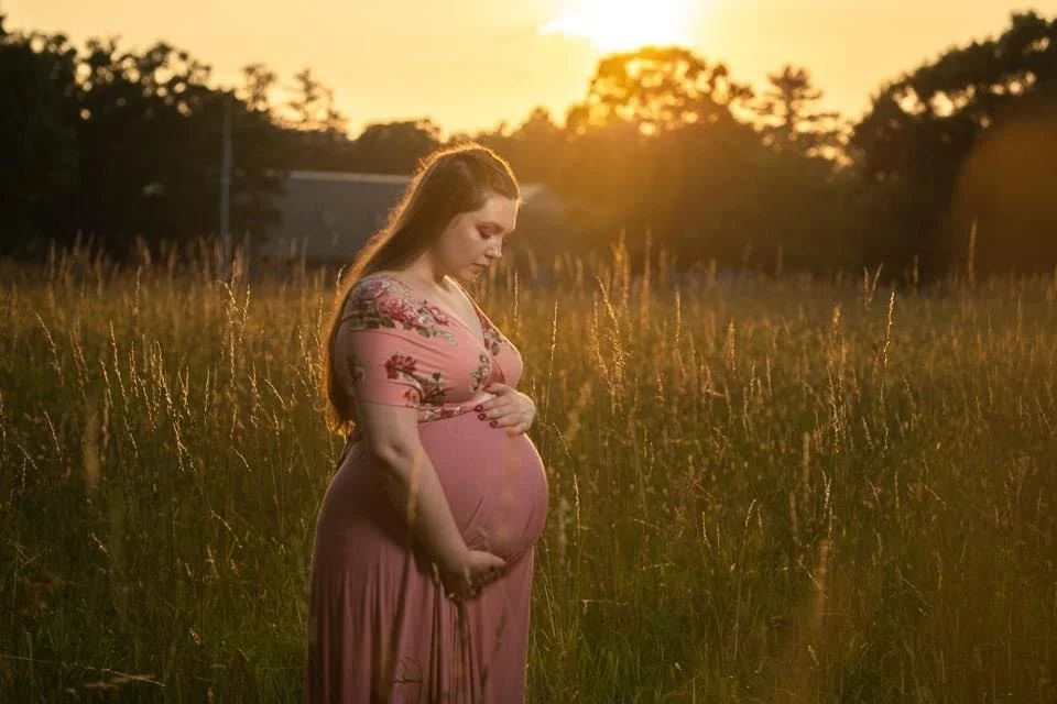 A pregnant woman in A maternity photo with a pink dress with floral embroidery stands in a field at sunset, looking down and touching her belly.