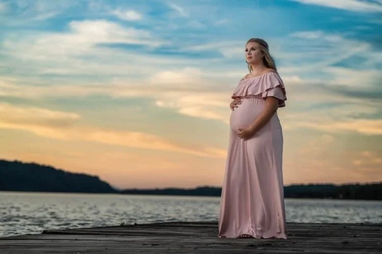 A maternity photo of A pregnant woman in a pink dress standing on a dock by a lake during sunset in the hudson valley.