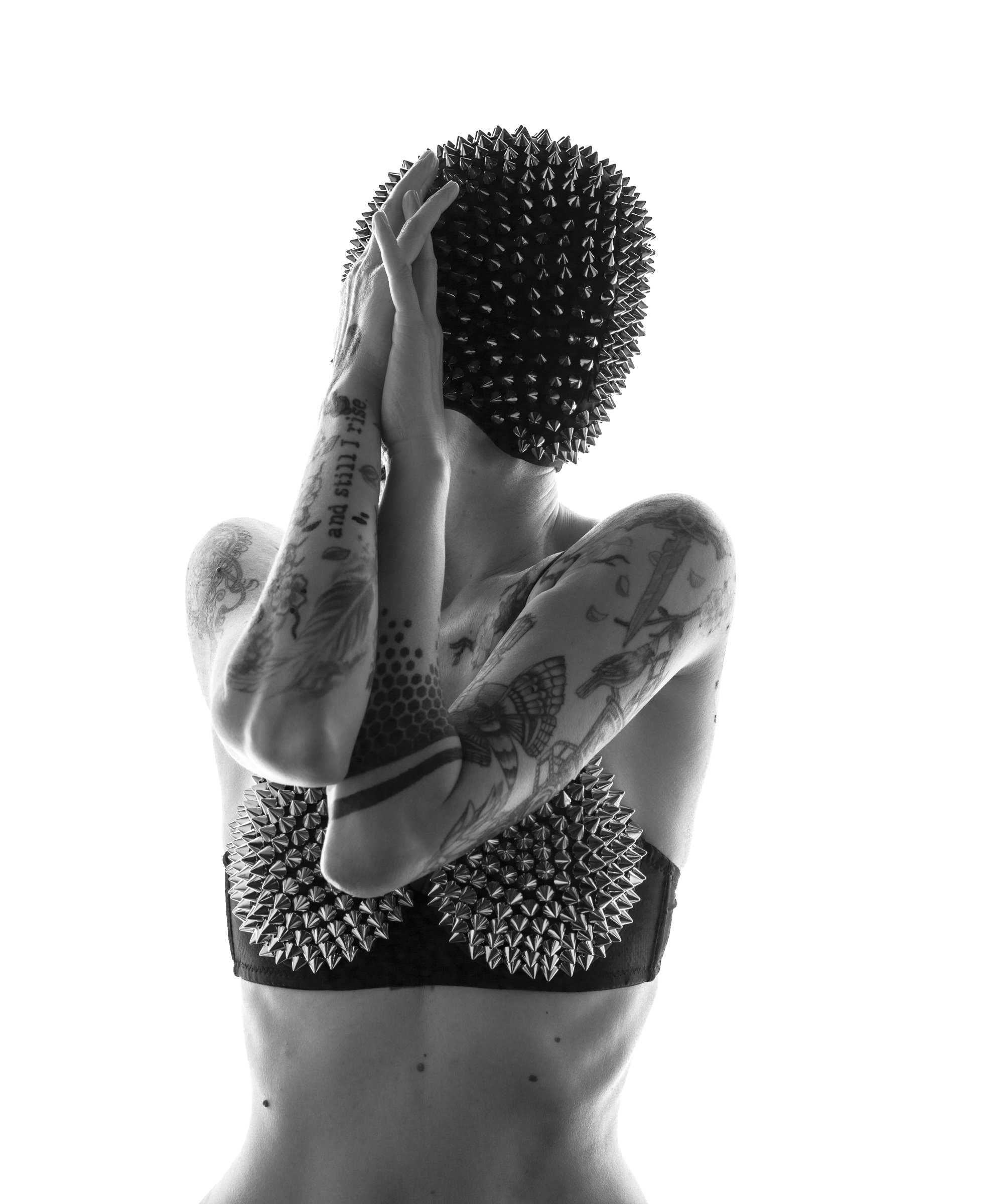 A person with tattoos covering their arms and torso, holding their head with one hand. Their face is obscured by a large, spiked, spherical object. The image is in black and white.