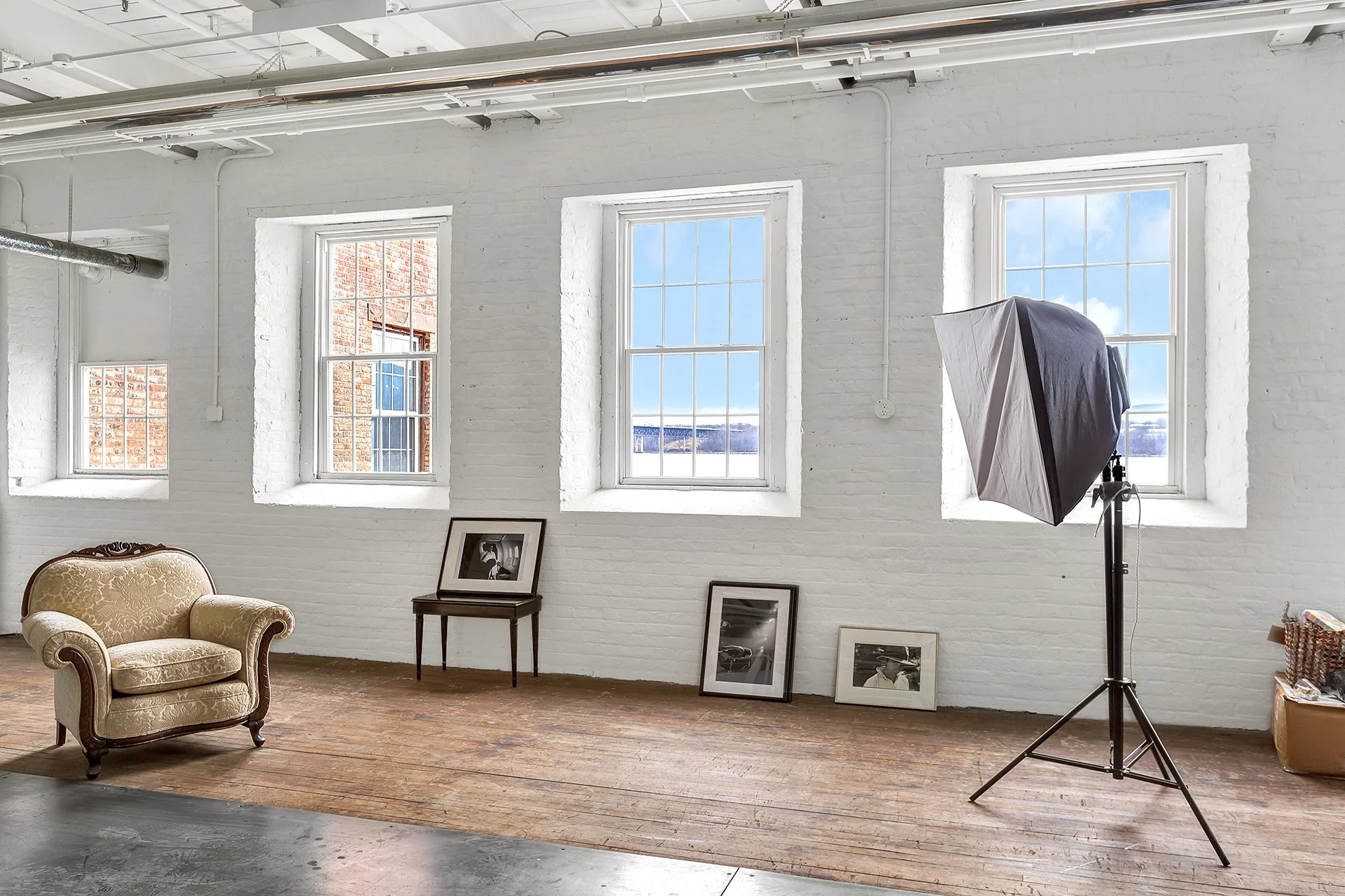 Interior of a photography studio with three large windows, a vintage armchair, framed photographs on the floor, and professional studio lighting equipment set up.