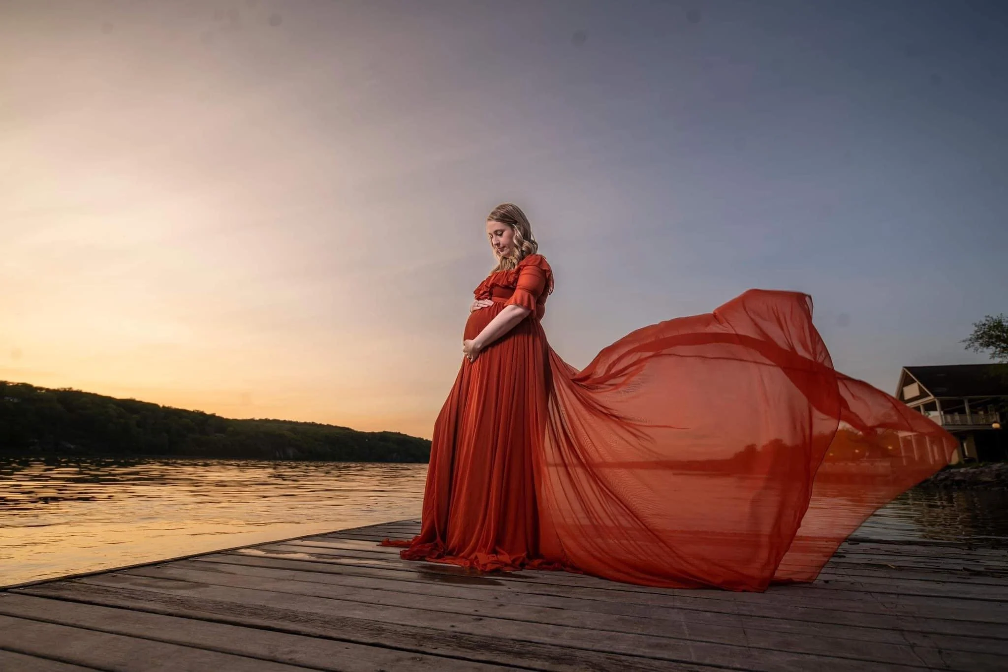 A maternity photo of a Pregnant woman in a flowing red dress standing on a wooden dock by a calm river during sunset.