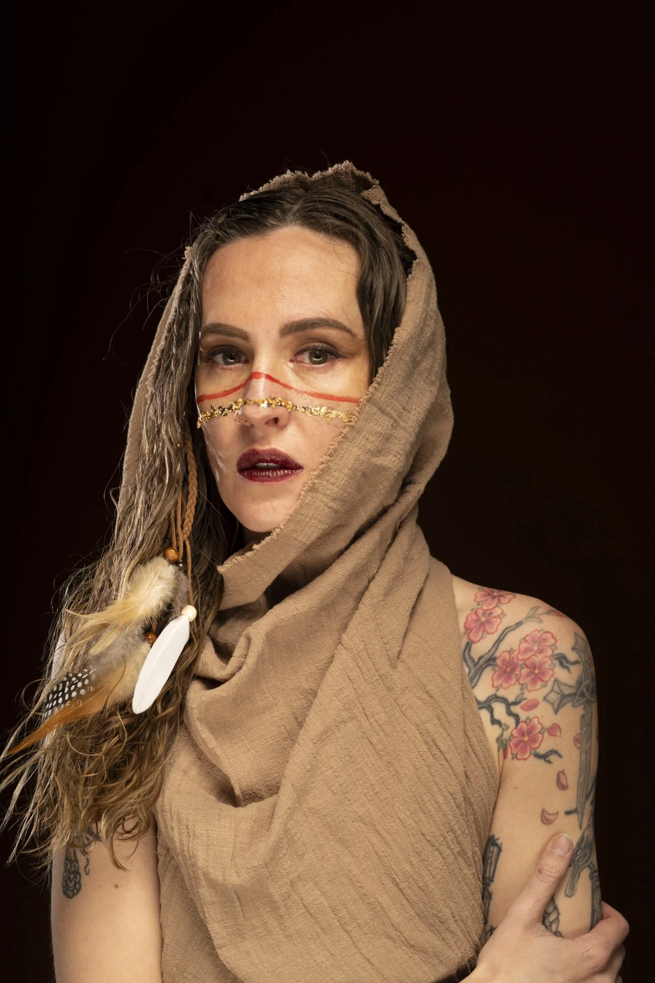 A woman with tattoos on her arm and a shoulder that has pink flowers, wearing a beige garment and a hood. She has long, wavy hair with beads and feathers, and face paint with red and gold elements. She has dark lipstick and makeup, posing against a b