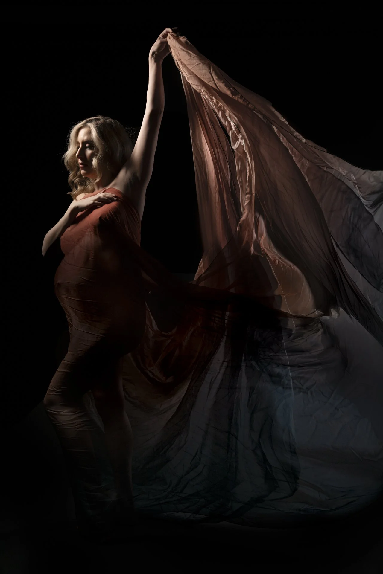 A woman with wavy blonde hair posing with one arm raised, holding a flowing piece of fabric, against a black background.