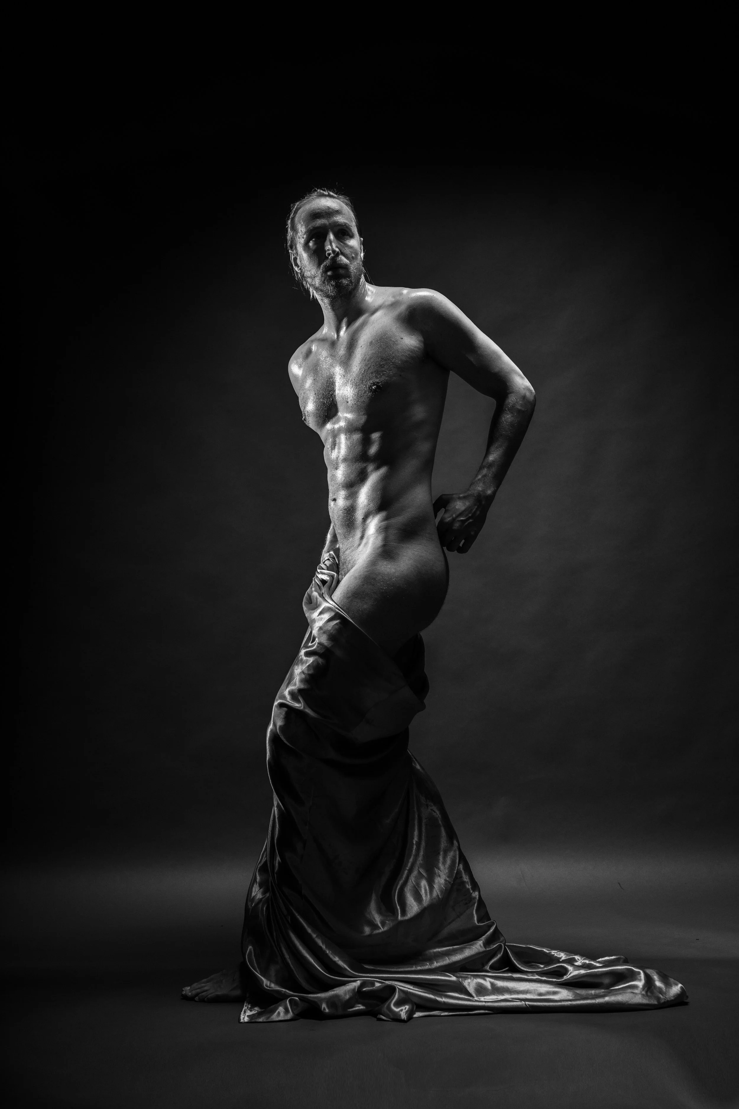 Black and white photograph of a shirtless man with a beard and long hair, kneeling on the ground with a satin sheet draped around his lower body. The background is dark and plain.