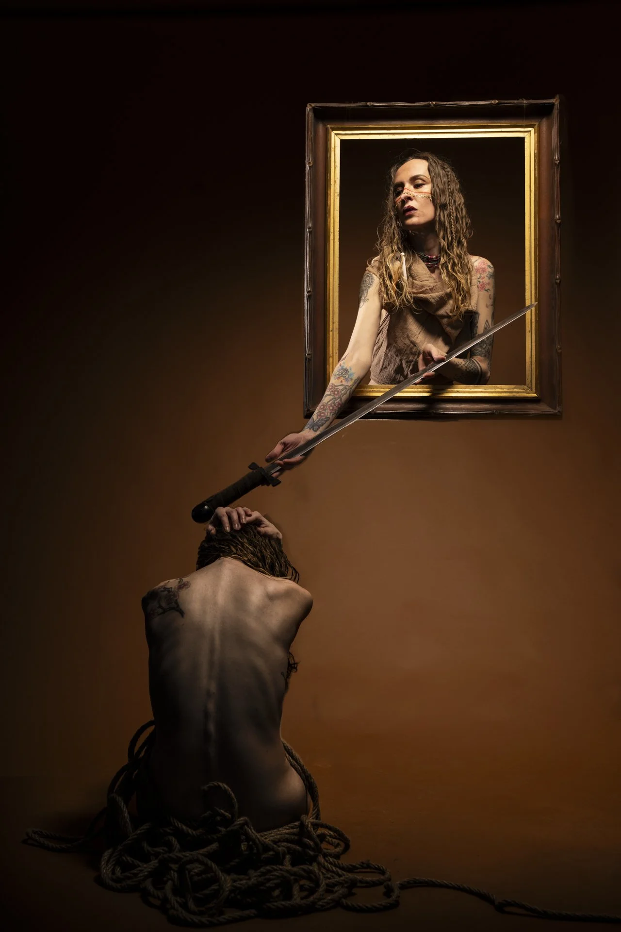 A woman with tattoos looking out of a picture frame on the wall, holding a sword. A man with long hair, facing away, is kneeling with his head bowed and hands supporting his head. The scene is dramatic with a dark background and a pile of ropes at th