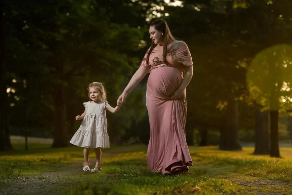 A maternity photo of A pregnant woman in a pink dress holding the hand of a young girl in a white dress in a park with trees during sunset.