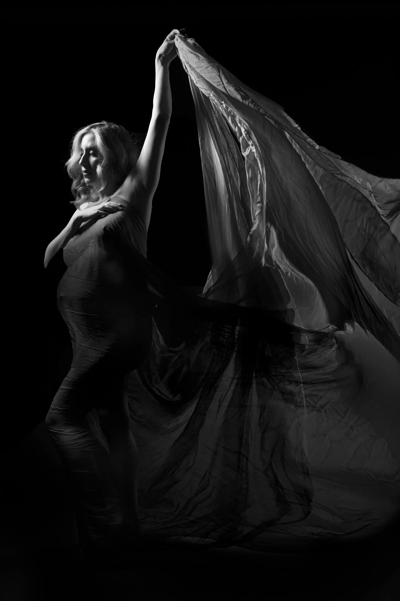 A woman with blonde hair in a strapless dress, holding a flowing sheer fabric in a black and white photo with dramatic lighting.