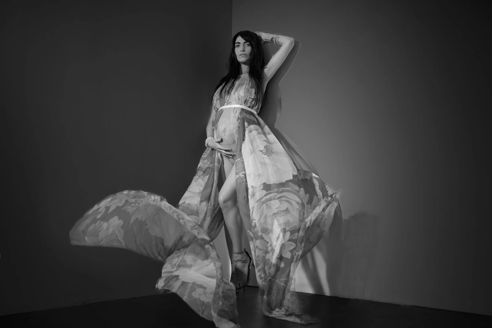 A black and white Maternity photo of a woman with dark hair wearing a floral dress with a slit, standing against a plain wall. She has one arm raised behind her head and is holding the dress open with the other hand.