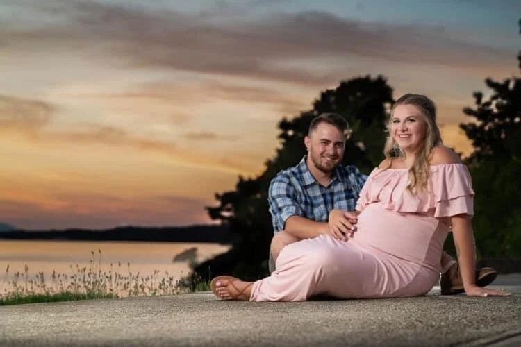 A maternity photo of A pregnant woman and a man sitting on the ground by a lake during sunset, smiling at the camera.