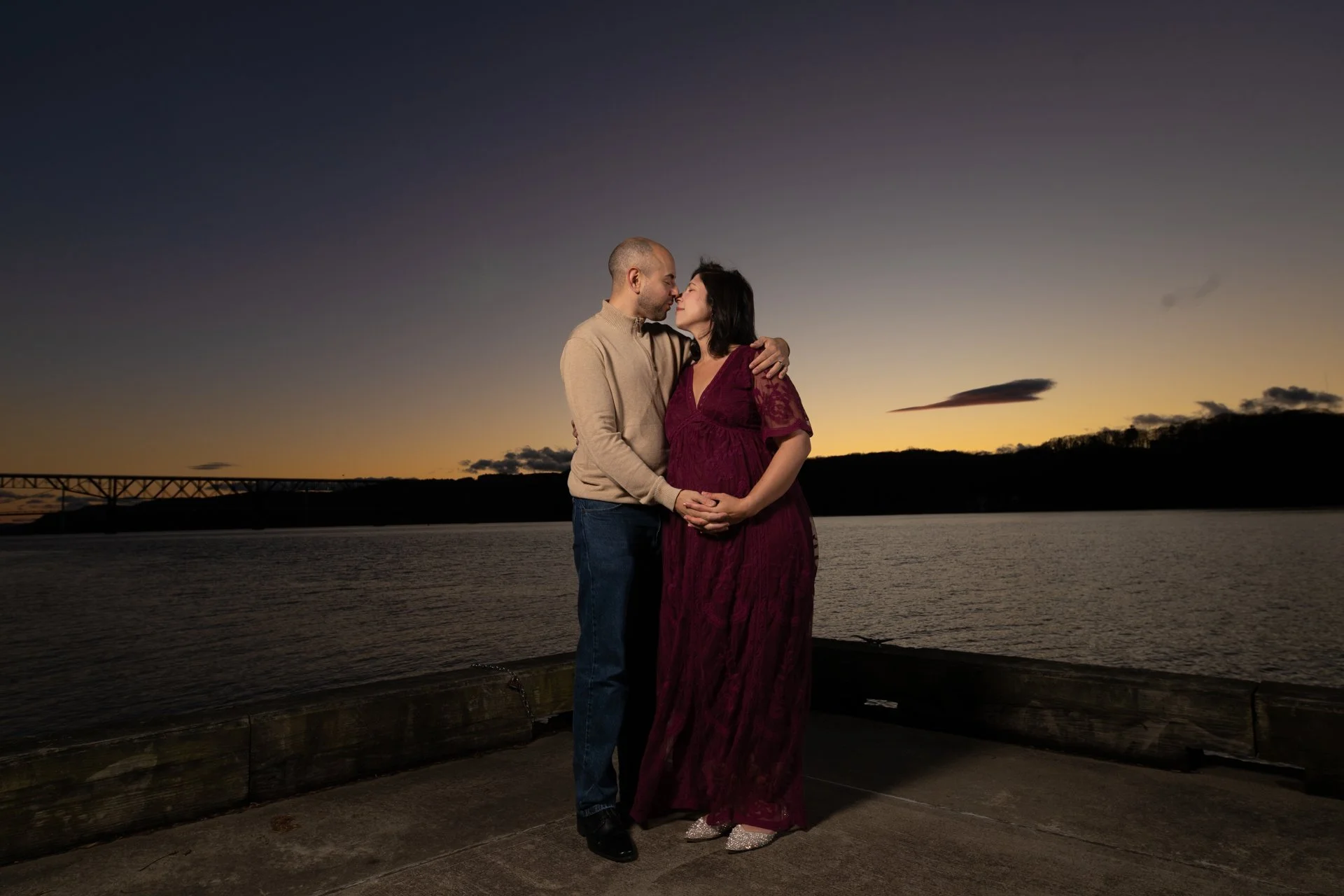A couple stands close together by a river at sunset, gazing into each other's eyes and holding hands. The man has a beard and is wearing a beige sweater, while the pregnant woman is in a burgundy lace dress. The sky is darkening with a scenic view of