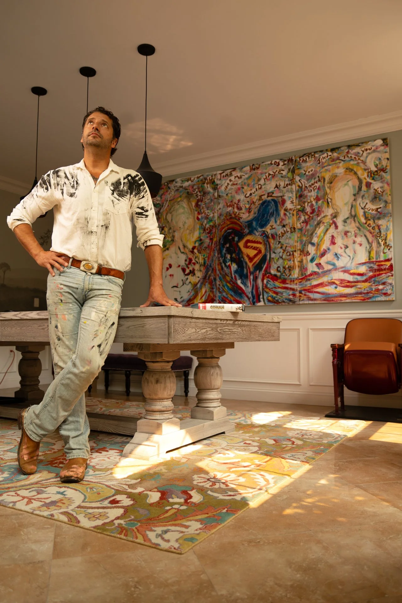 A man standing in a room with his left hand on his hip, wearing a white shirt with black paint splashes, jeans with paint marks, and brown boots. Behind him, there is a large colorful abstract painting on the wall, a wooden table with a book on top, 
