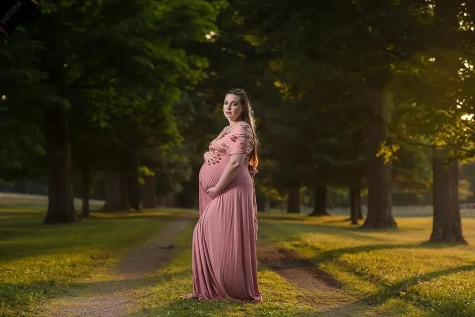Pregnant woman in a pink dress standing on a grassy path in a park with tall trees at sunset. A maternity photo.