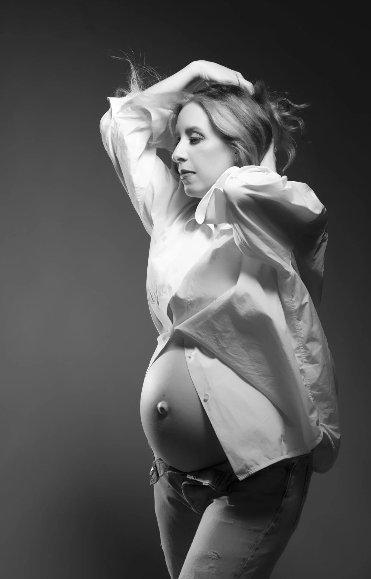a maternity photo in the hudson valley Black and white photo of a woman with one arm raised and hand on her head, wearing an unbuttoned shirt exposing her belly and jeans.
