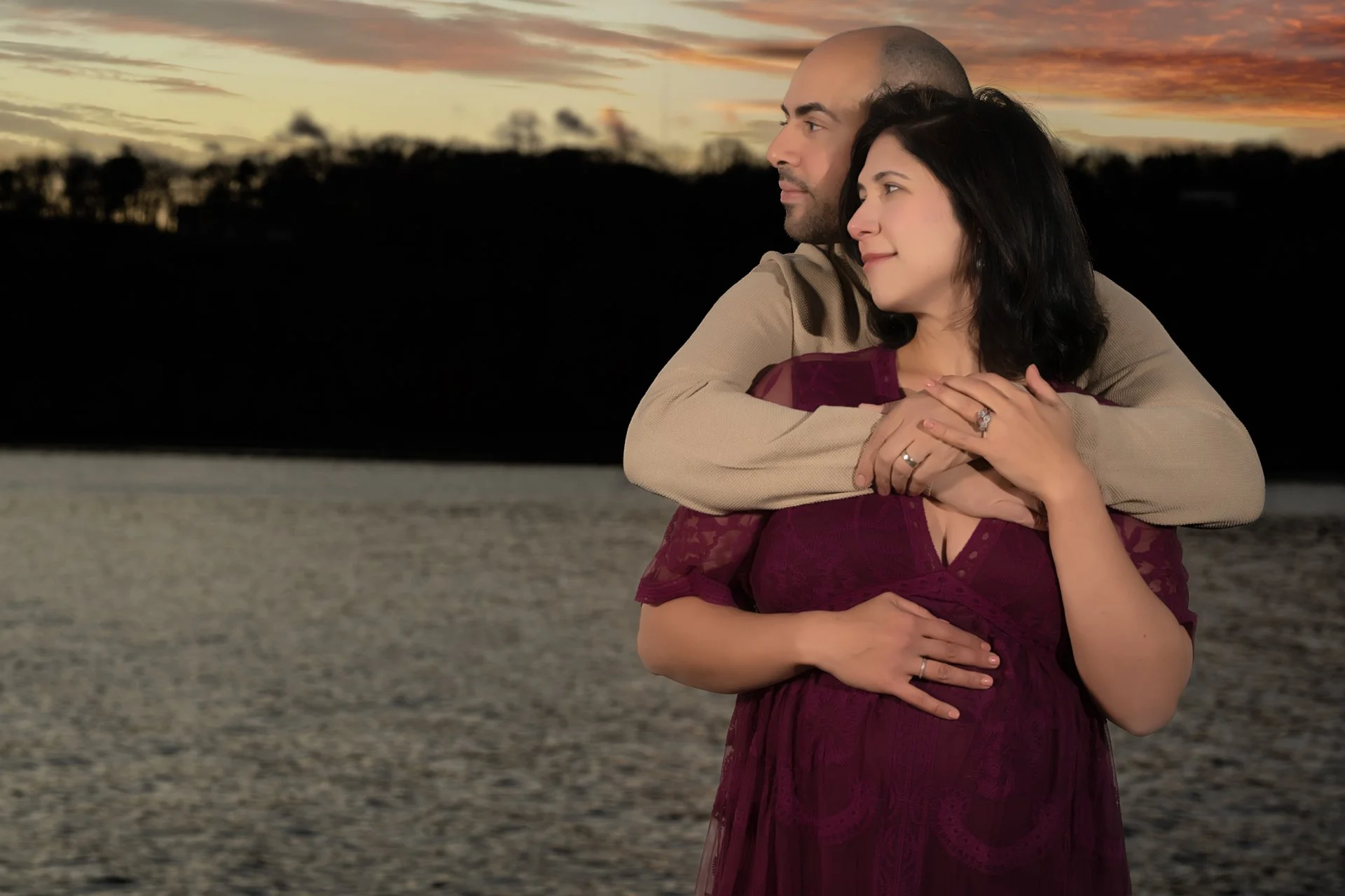 a maternity photo featuring A couple embracing near a the hudson river during sunset, with the man holding the woman from behind as they look into the distance.