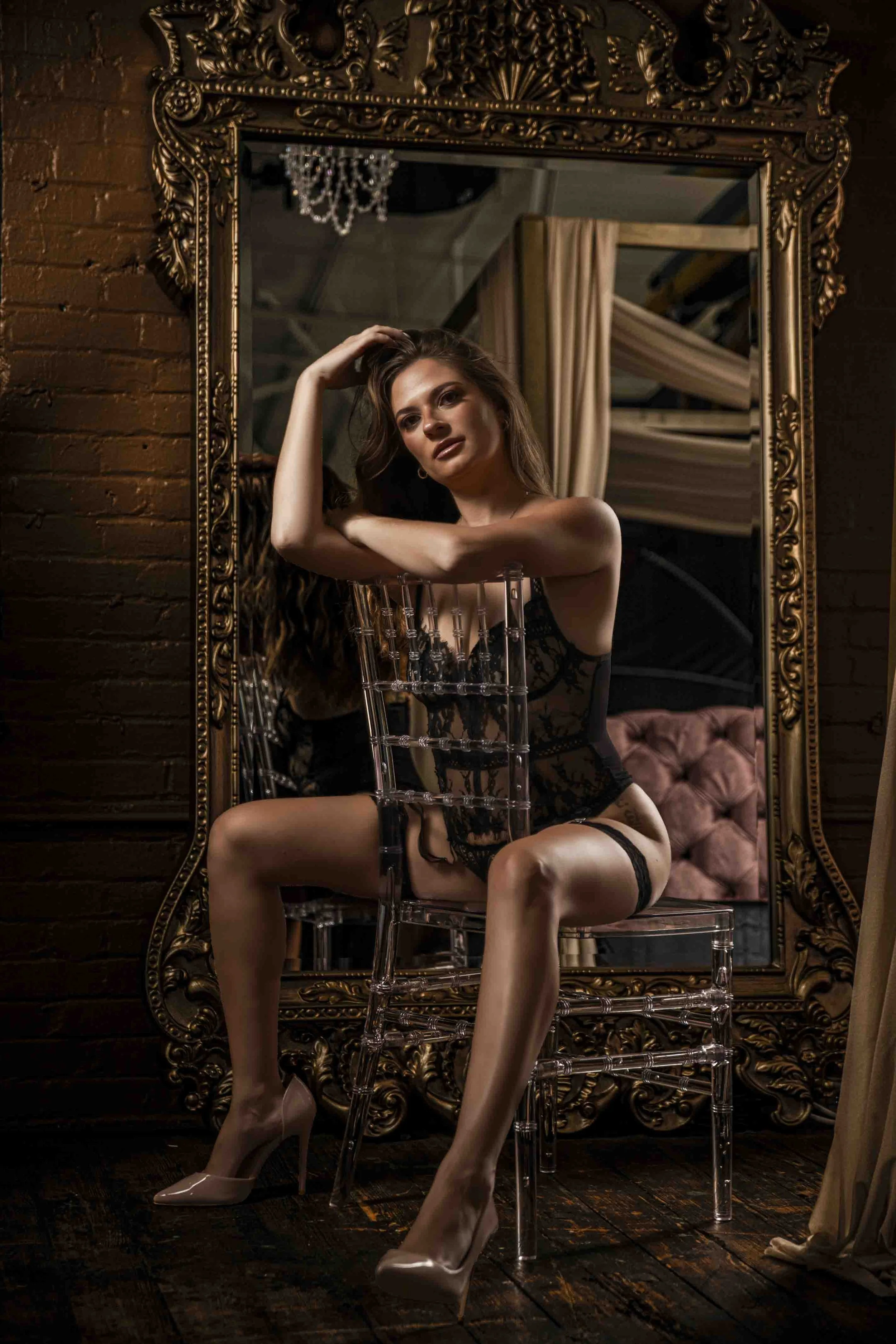 A woman with long brown hair sitting on a clear acrylic chair, posing in front of a large ornate gold mirror. She is wearing black lace lingerie, beige high heels, and thigh-high stockings. The setting appears to be a dimly lit, stylish room with dar