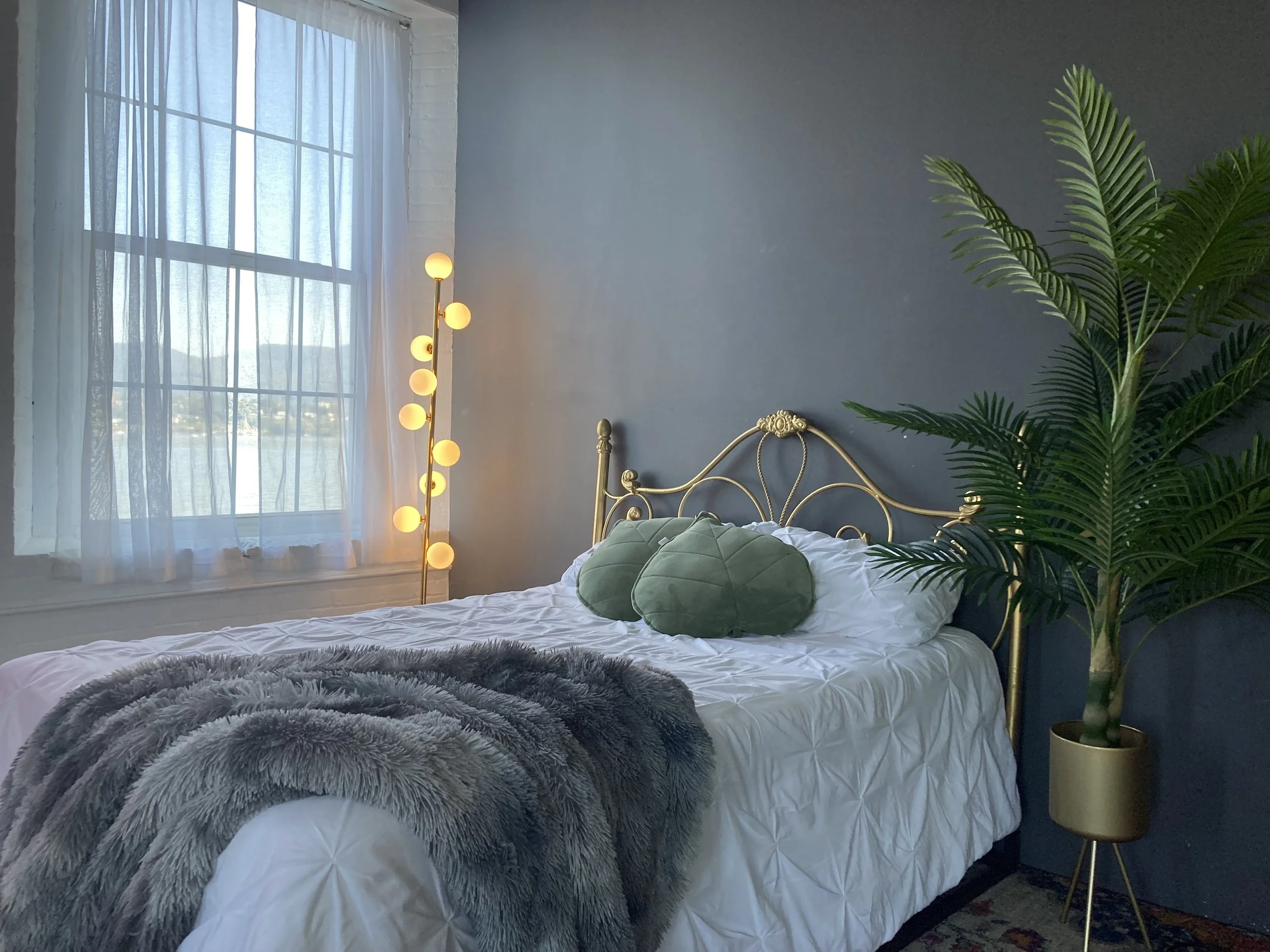 A bedroom with a large window covered by sheer curtains, a brass bed with a white quilt, a green velvet pillow, a fluffy gray throw, a tall plant in a pot, a gold floor lamp with round bulbs, and a blue wall.