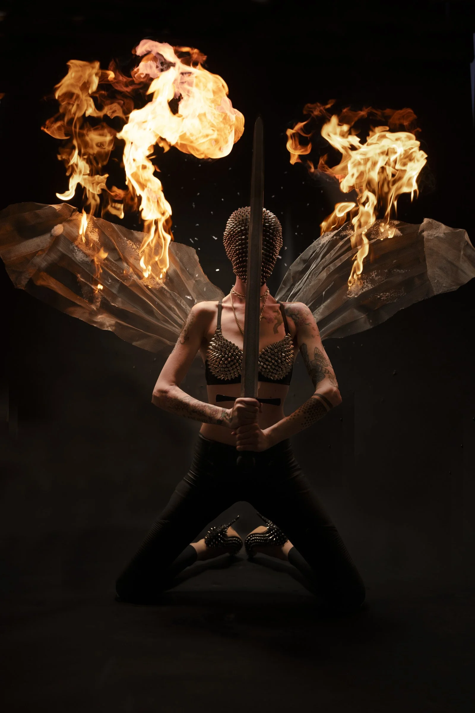 A person with tattoos, wearing a spiked bra, standing in a wide stance and holding a sword vertically in front of their face. They have large, wing-like structures made of reflective material and flames erupting from their head against a dark backgro
