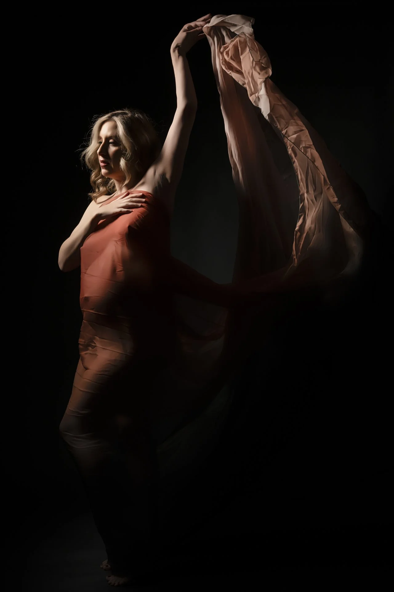 A woman with blonde hair in a strapless dress poses with one hand on her chest and the other raised, holding a flowing piece of fabric against a dark background.