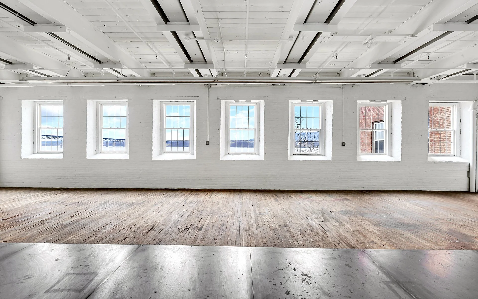 Empty industrial-style room with six windows, white painted brick walls, wooden flooring, and exposed ceiling beams and piping.