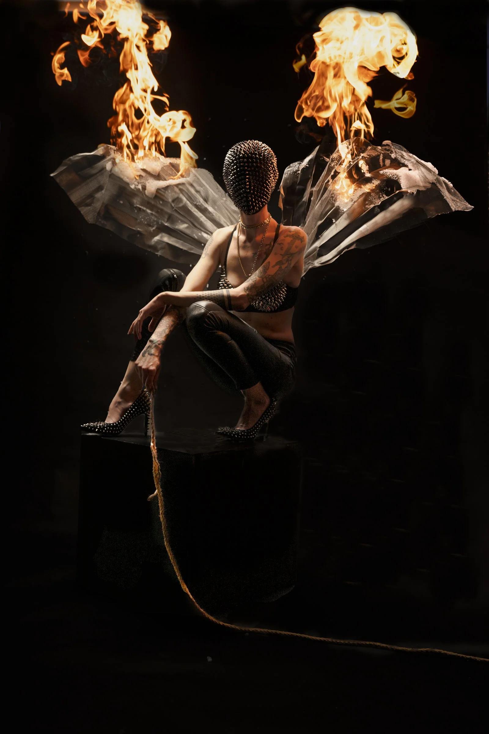 A person with tattoos, wearing a studded face mask, sitting with legs crossed, dressed in black leather pants and high heels, with large butterfly wings made of paper or fabric behind them, and flames rising from each wing.