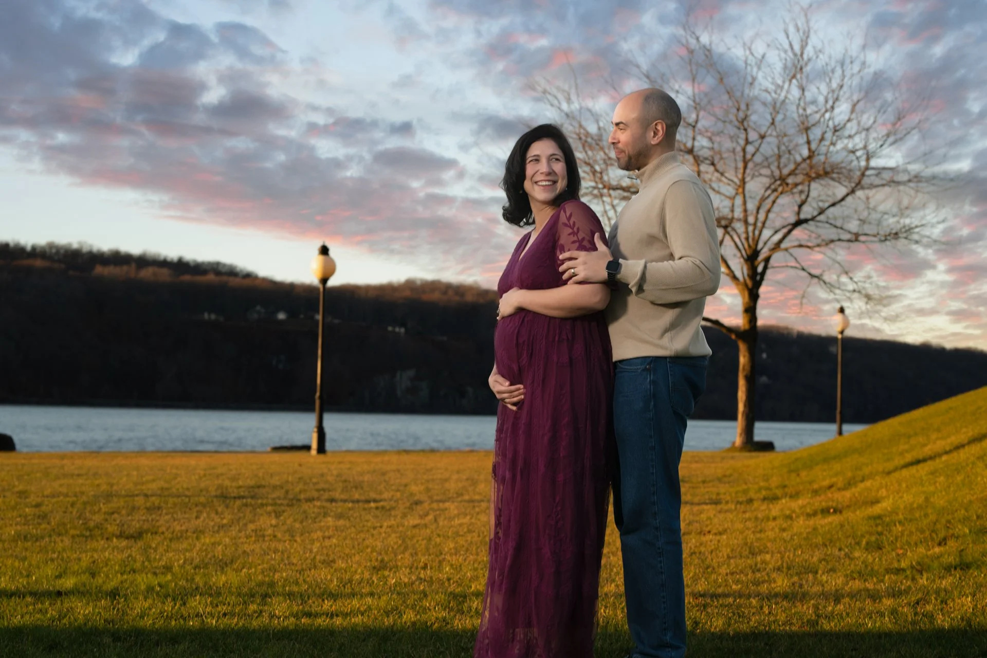 a maternity photo featuring A pregnant woman in a purple dress and a man in a beige sweater are standing outdoors at sunset near a lake, with trees and lamp posts in the background.