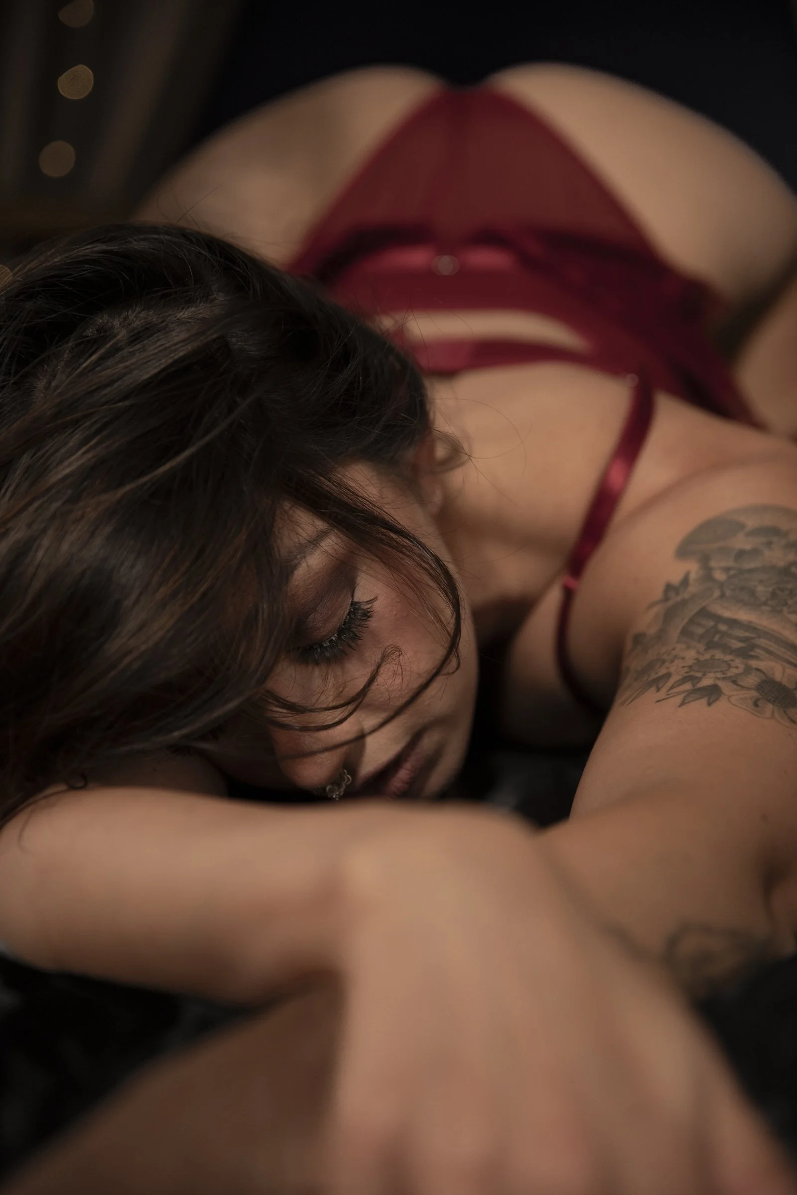A woman in red lingerie lying on her side with her head resting on her arm, showing tattoos on her arm and dark hair falling over her face.