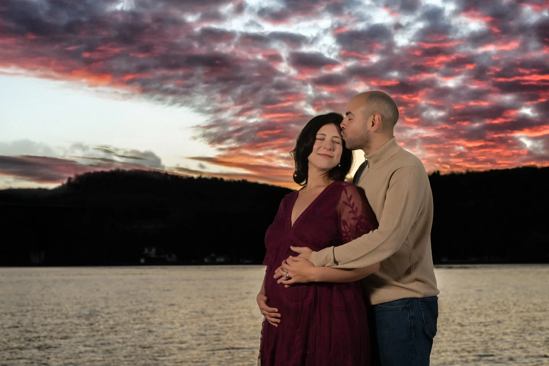 a maternity photo in the hudson valley featuring A couple standing by the water during sunset, with the man kissing the woman's temple, and the woman smiling with closed eyes.