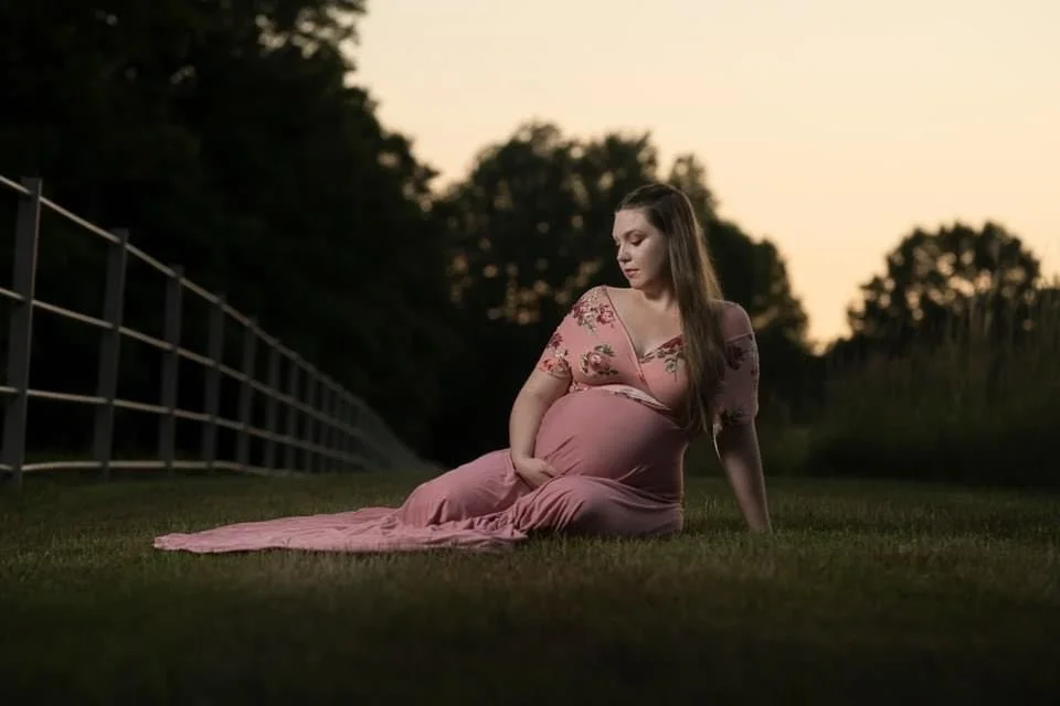 A pregnant woman in a pink floral dress sitting on the grass next to a fence during dusk, looking down at her belly, A maternity photo.