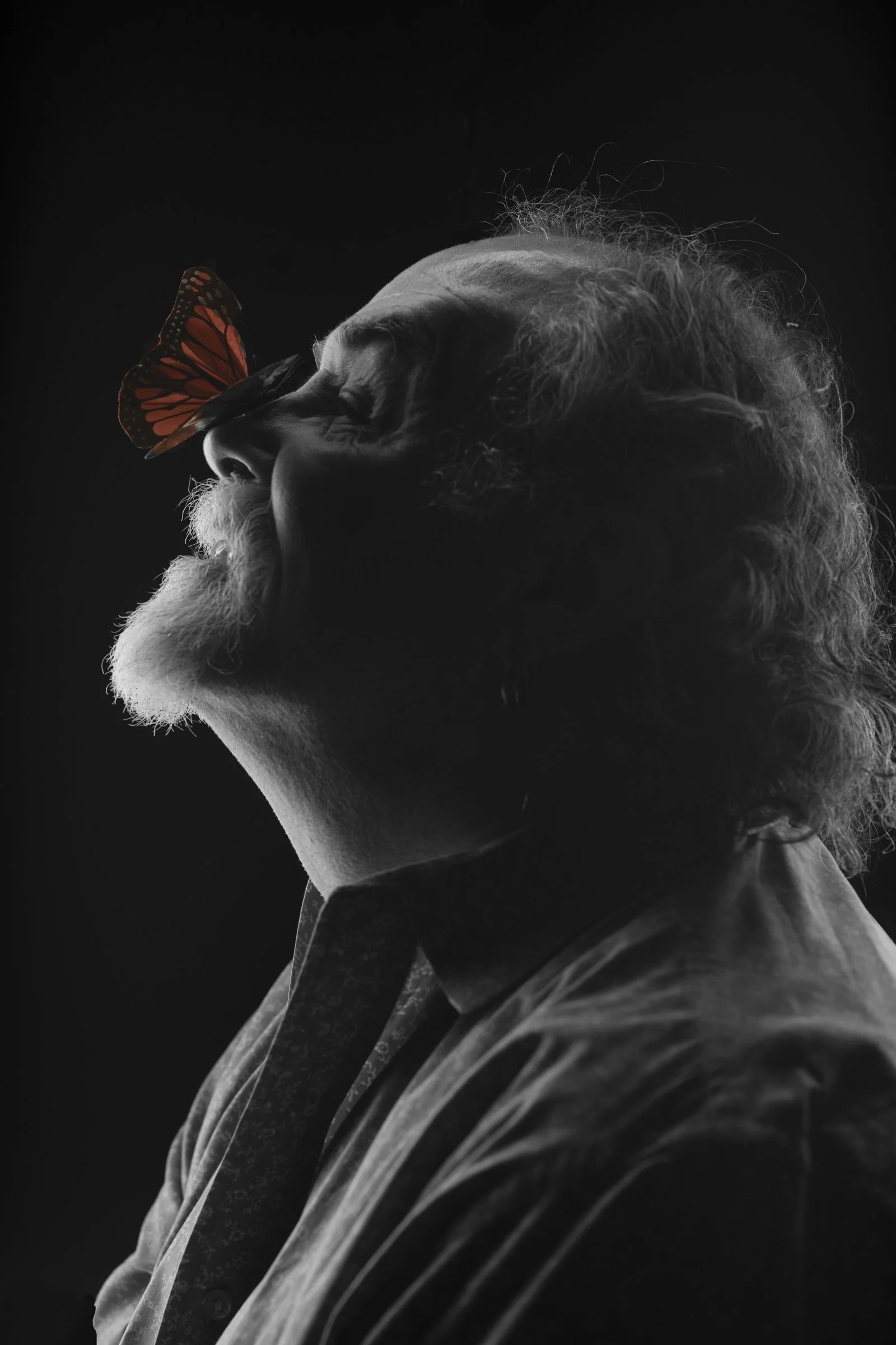 A black and white portrait of an elderly man with a full head of curly hair and a beard. A monarch butterfly is resting on the man's nose, and the man appears to be smiling with his eyes closed.
