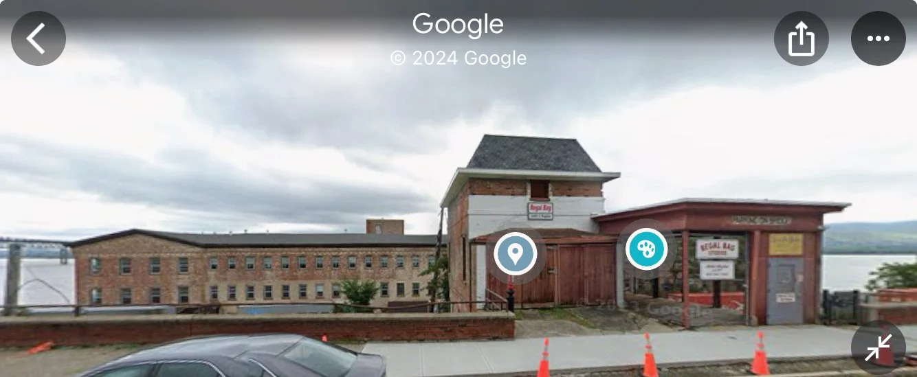 A brick building with a waterway view, a small entrance with signs, and orange safety cones on the sidewalk.