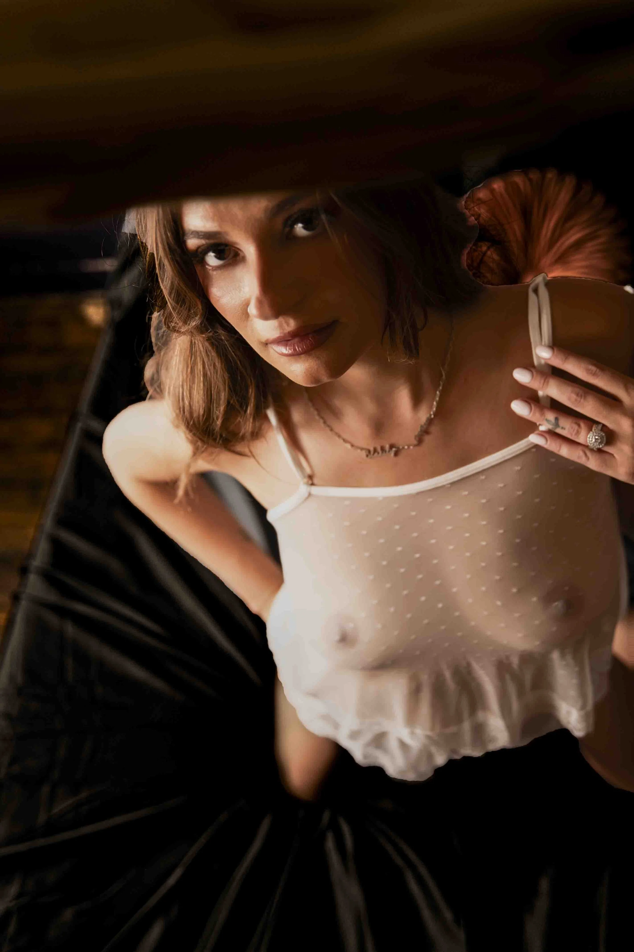 A woman with light brown hair, wearing a beige polka dot top and jewelry, looking up through a gap in dark furniture.