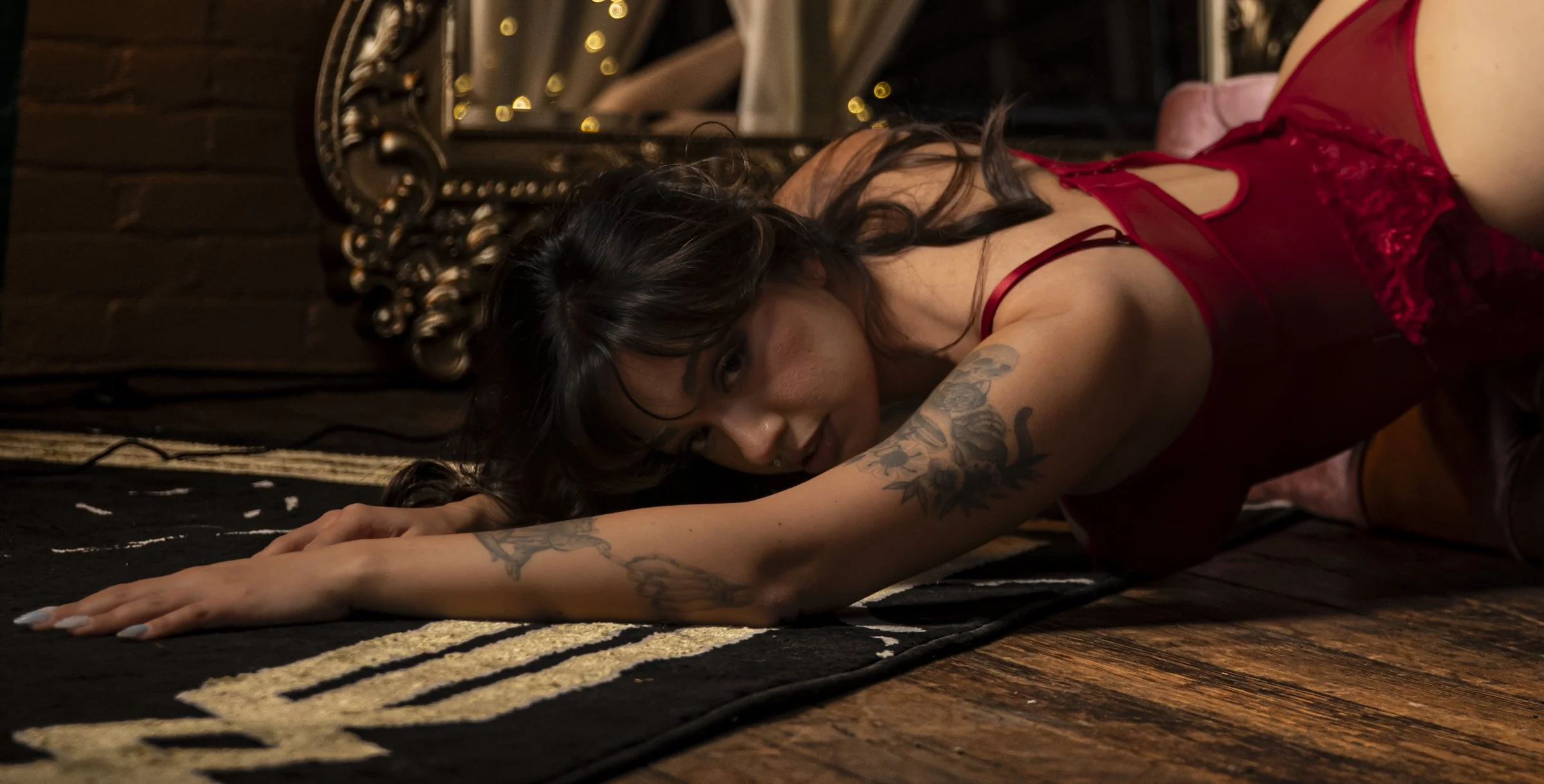 A woman with dark hair and tattoos lying on a rug in a dimly lit room, wearing red lingerie.