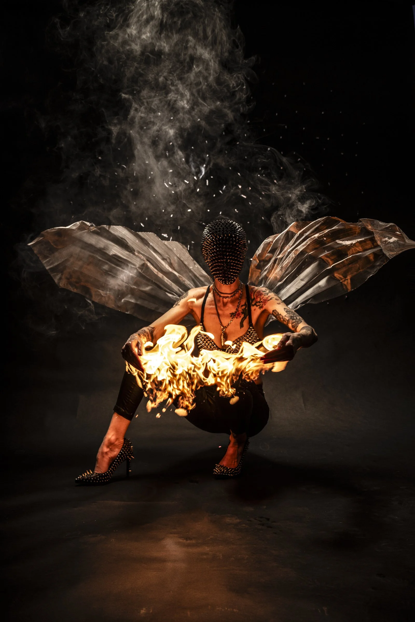 A person with a studded mask and tattoos, holding fire, kneeling with one leg extended and flames around their arms, with metallic wings on their back, against a dark background.