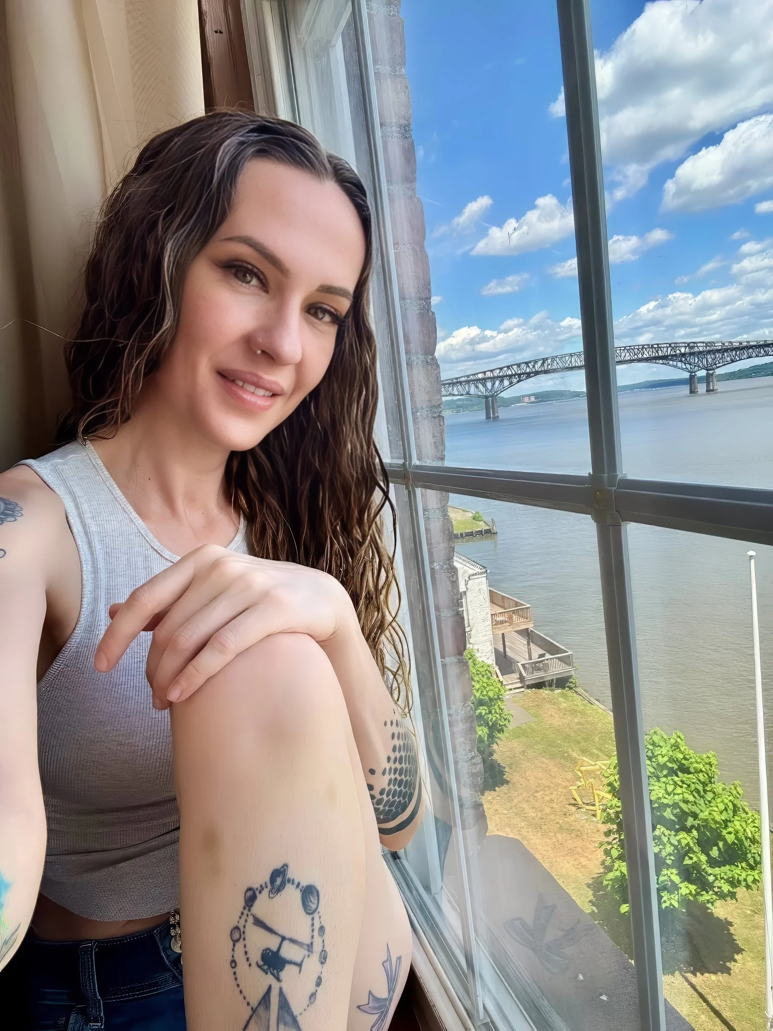 A young woman with long, wavy brown hair and light makeup sitting by a window on a bright, sunny day. She is wearing a gray sleeveless top and has tattoos on her left arm and side. Behind her, the view shows a river, a bridge, and a partly cloudy sky