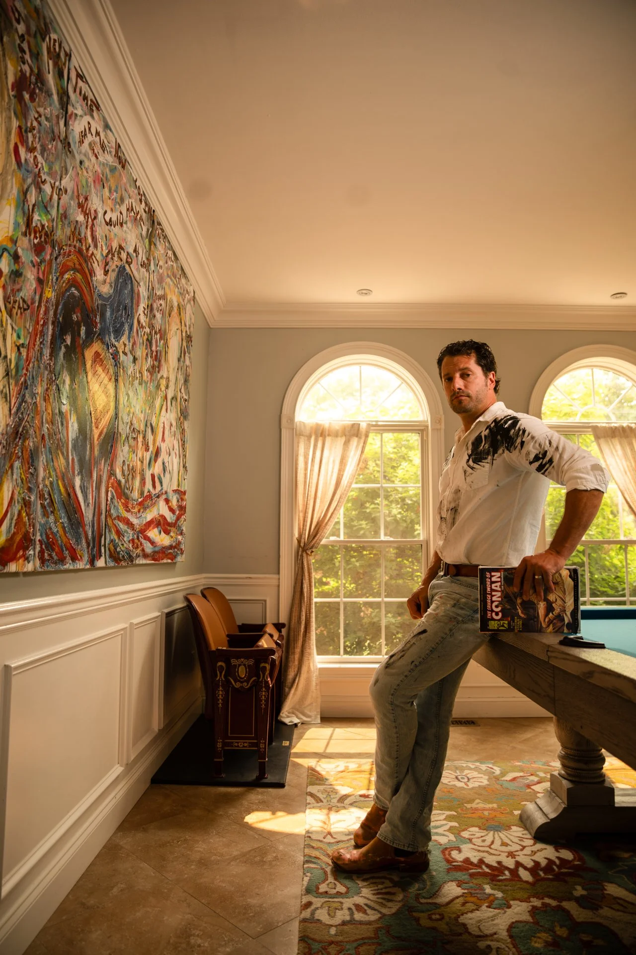 A man in a white shirt with black paint marks and light-colored jeans stands in a brightly lit room holding a book. He leans against a sturdy wooden table with floral-patterned fabric underneath. Artistic paintings hang on the wall, and large windows