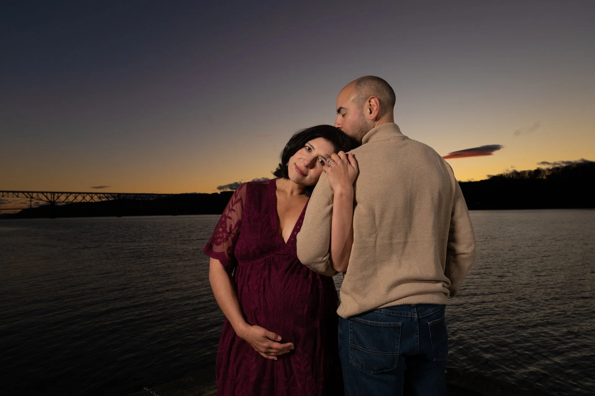 a maternity photo in the hudson valley featuring A couple standing by the water at sunset, with the woman resting her head on the man's shoulder and holding her pregnant belly.