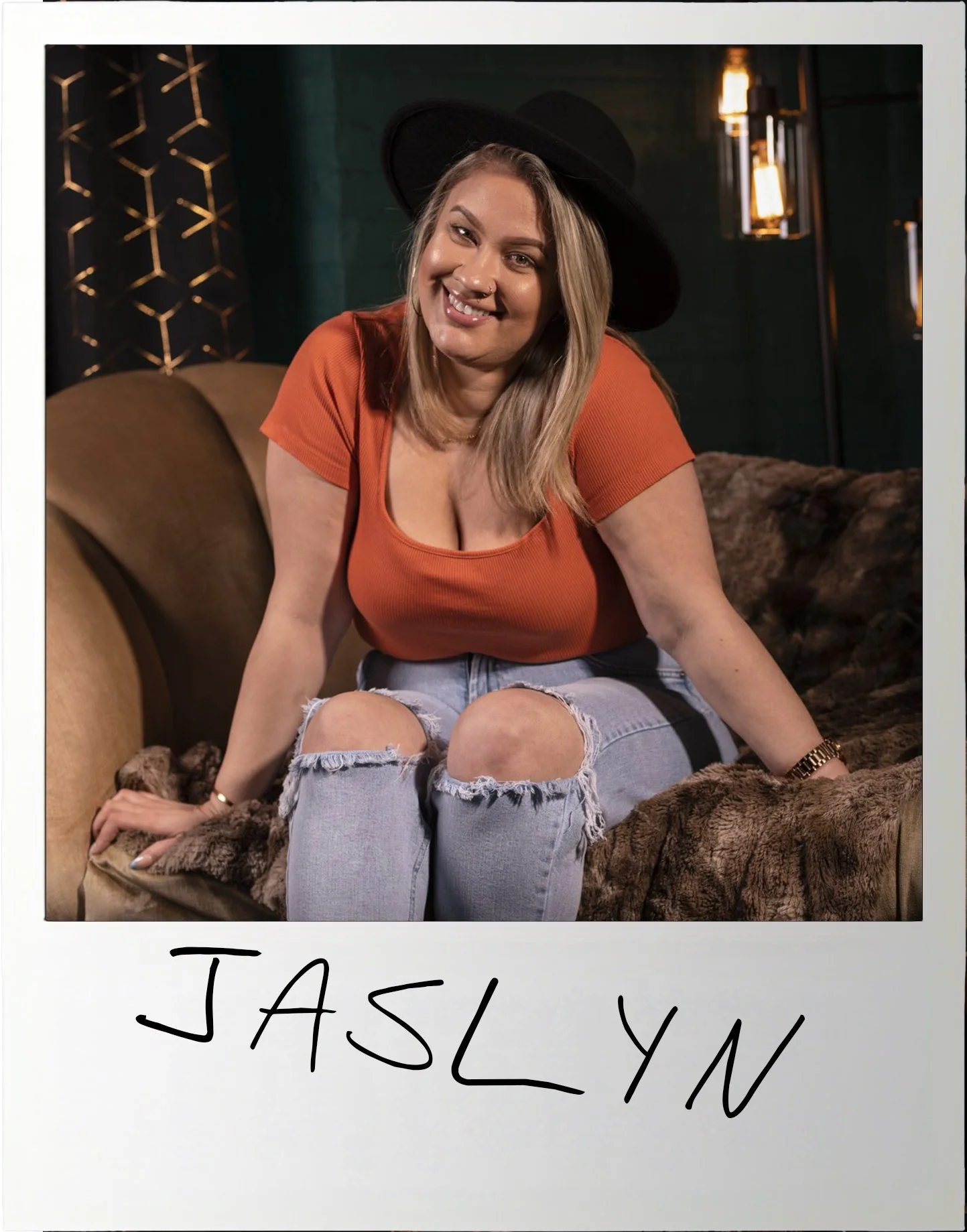 A woman sitting on a brown furry blanket, wearing a black wide-brimmed hat, an orange top, and ripped jeans, smiling at the camera in a cozy, dimly lit room with modern wall decor and warm lighting.