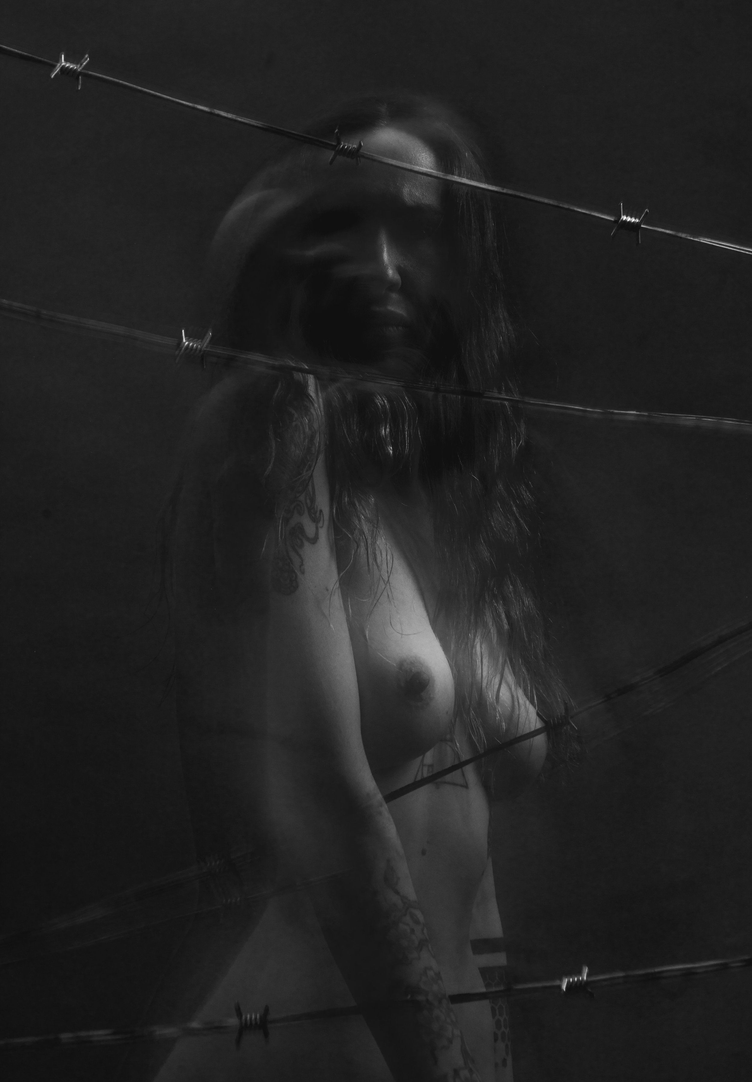Black and white photo of a woman with long, curly hair, wearing a hat, posing behind a glass surface with barbed wire.