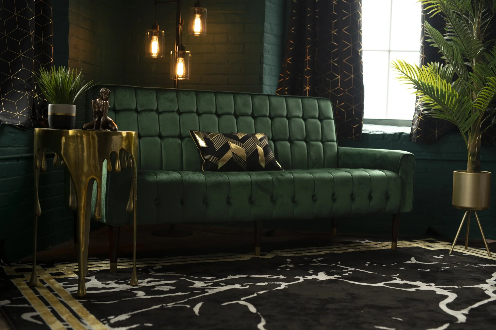 A stylish living room corner featuring a green velvet tufted sofa with a gold and black geometric patterned pillow. Next to it, a metallic gold side table holding a potted plant and a decorative figurine. Behind, a large window with black curtains wi