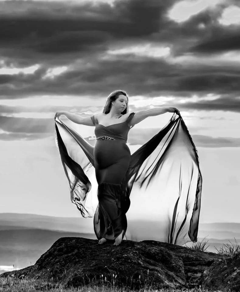 A woman standing on a rock outdoors, holding flowing fabric in each hand, with a mountainous landscape and cloudy sky in the background.