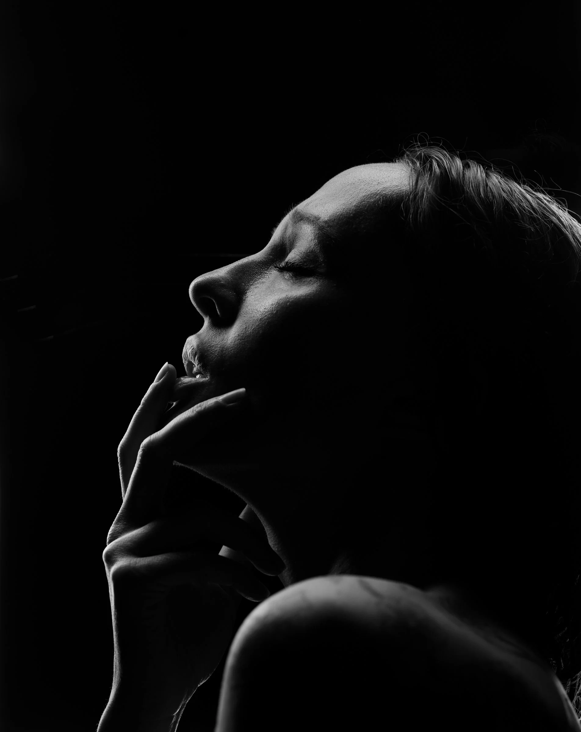 A black and white photo of a woman with closed eyes, holding her hand near her face, with dramatic lighting highlighting her facial features and hair.