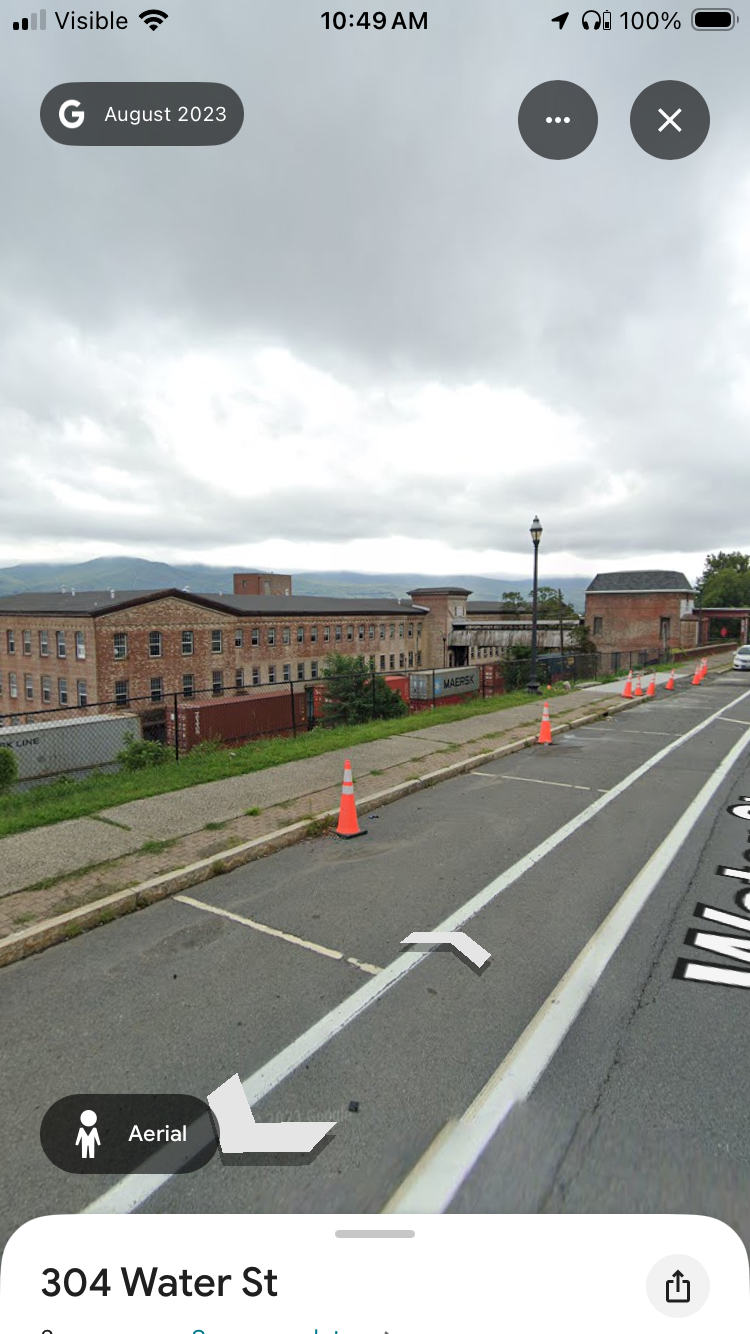 Street view with parking spaces marked on the side, orange traffic cones, a fence, a brick building, a lamppost, and overcast skies.