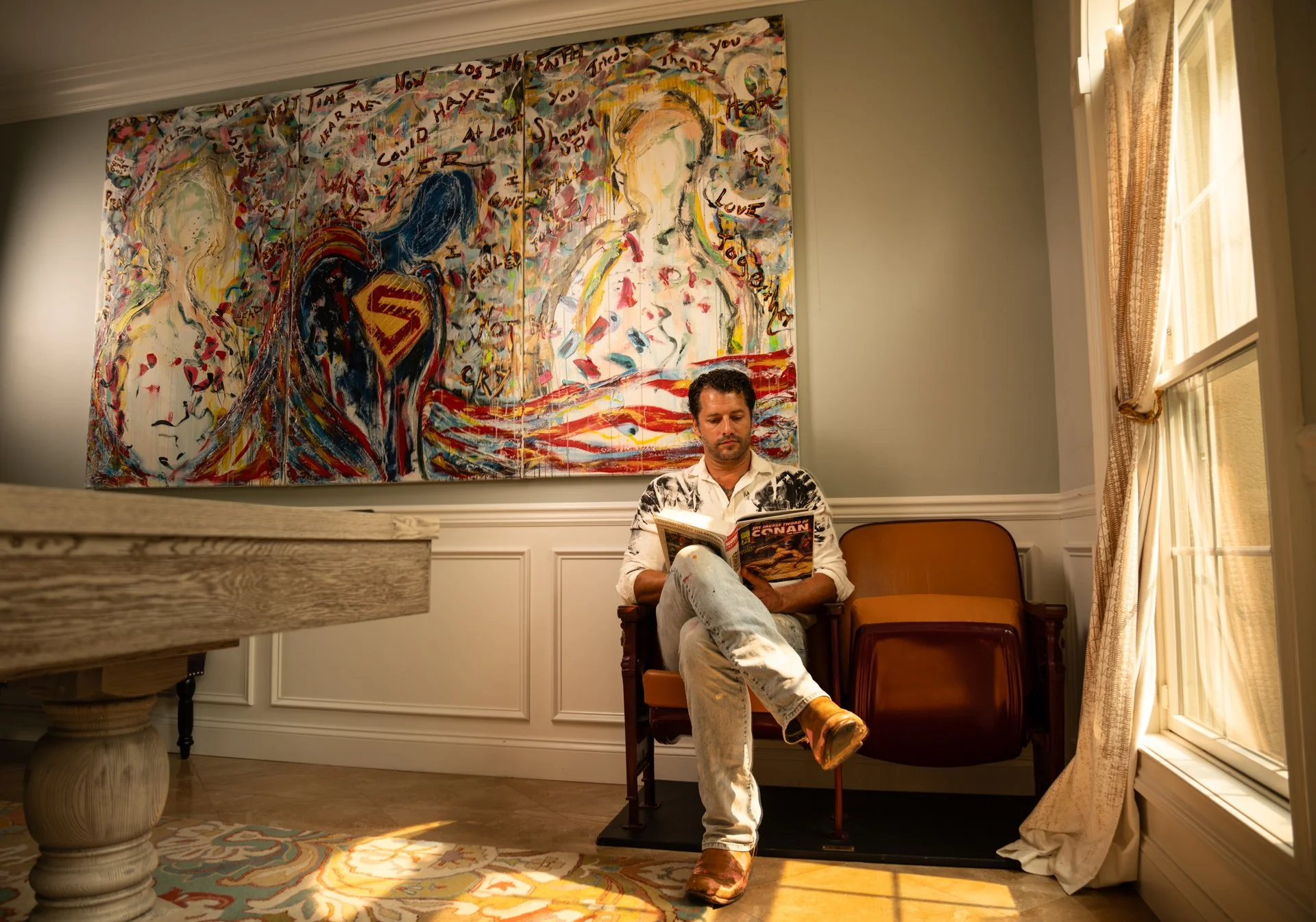 A fine art image of a man sitting in a chair reading a Conan magazine in a warmly lit room with a large abstract painting on the wall behind him. The room has cream-colored walls, a patterned area rug, and large windows with curtains.