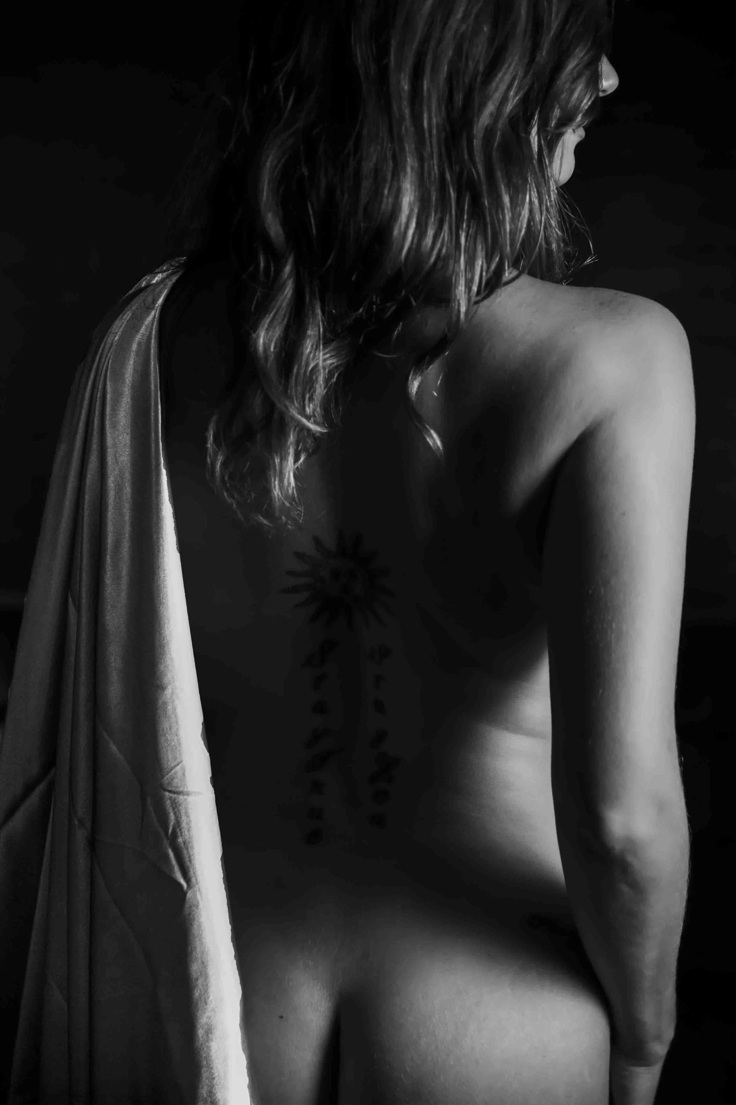 Black and white photo of a woman with wavy hair, turned sideways, showing her shoulder and back, with a tattoo of a sun on her shoulder blade, and a fabric draped over her left shoulder.