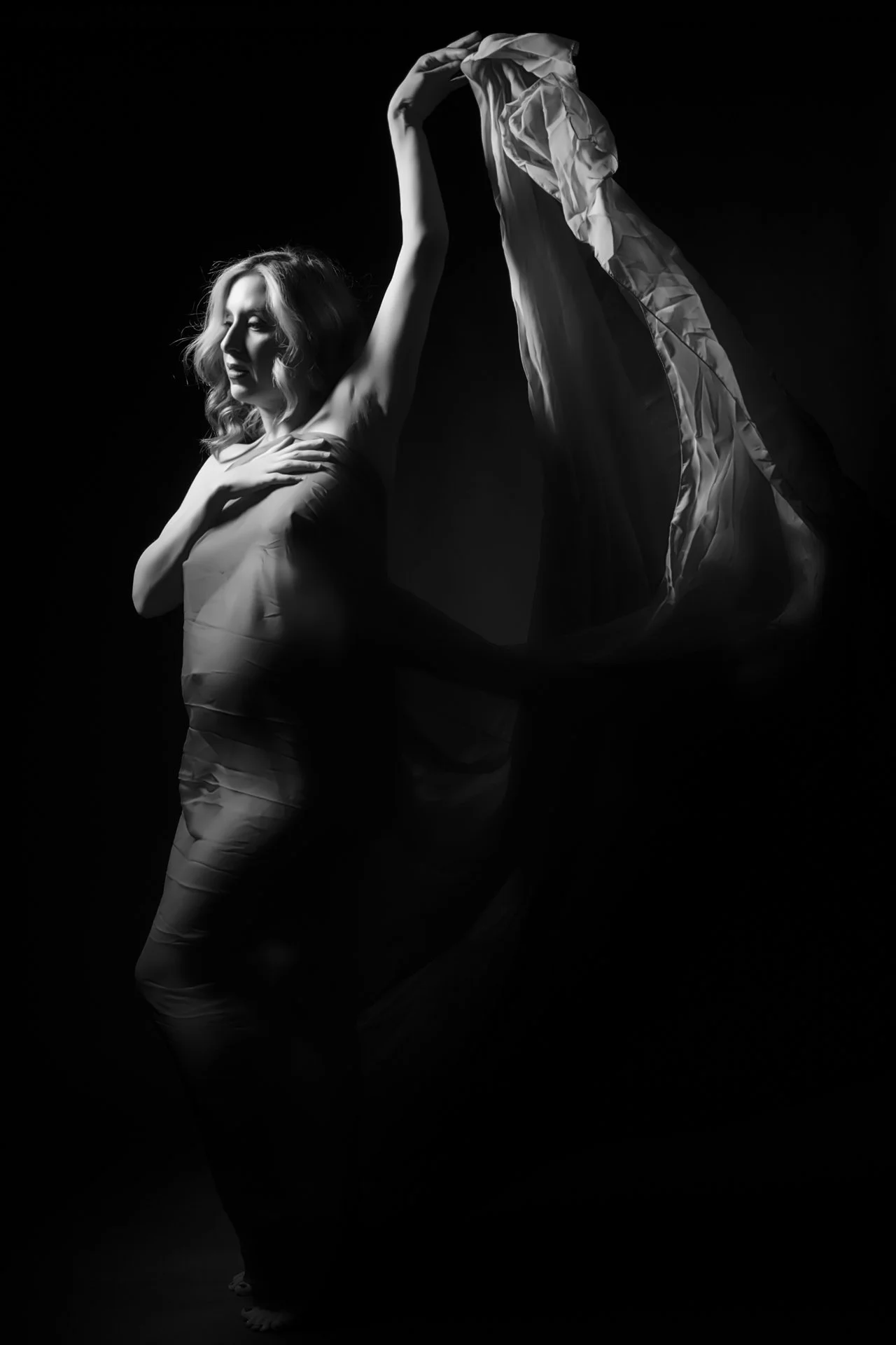 A black and white photo of a woman with wavy hair, holding a fabric or cloth above her head, with one hand on her chest and the other raised. The background is dark, creating a dramatic effect.
